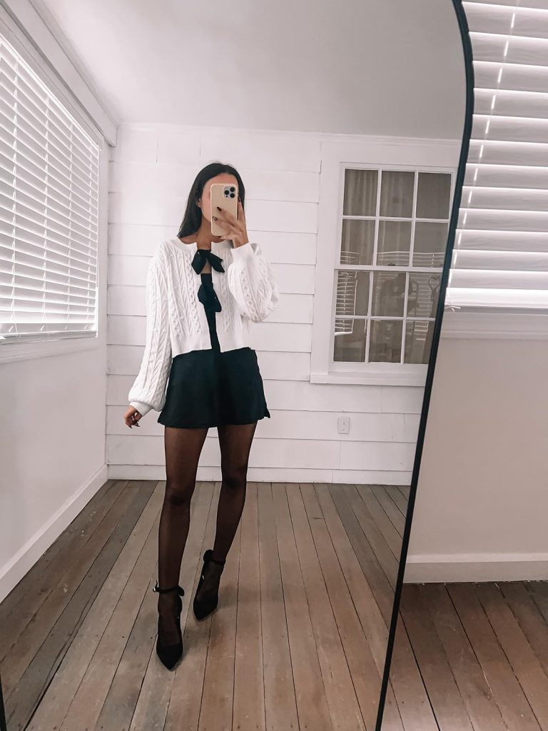 white cropped knit cardigan with black ties, a black mini dress, tights, and black heels